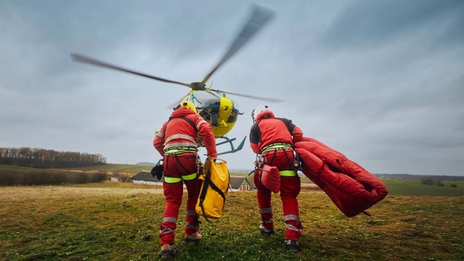 Two emergency responders carry equipment toward a waiting helicopter on a grassy field under a cloudy sky.