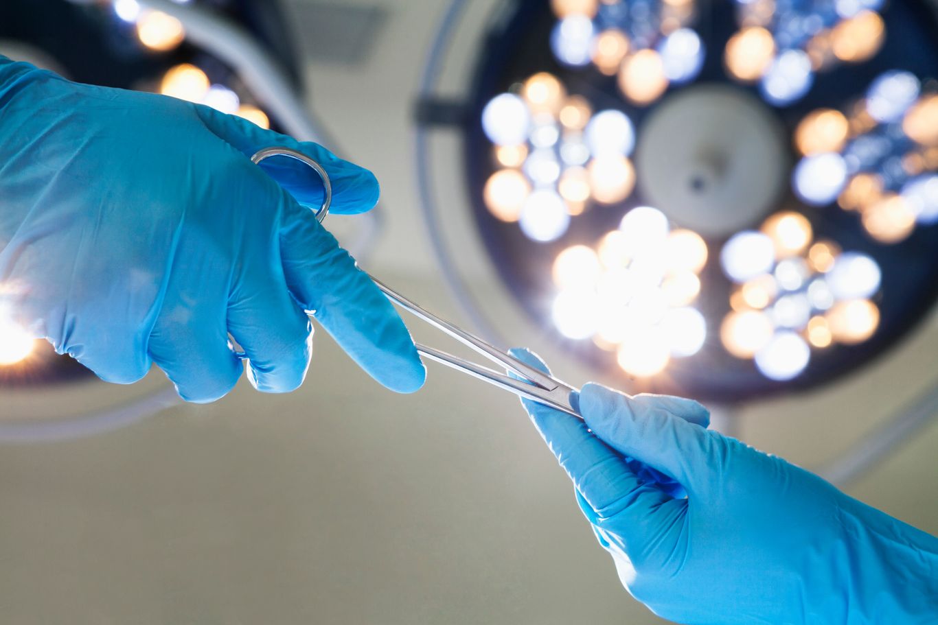 Two gloved hands exchange surgical instruments under operating room lights.