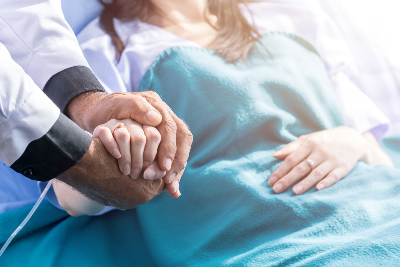 A doctor holds a patient's hand in a hospital bed, offering comfort and support. The patient is covered with a blue blanket.