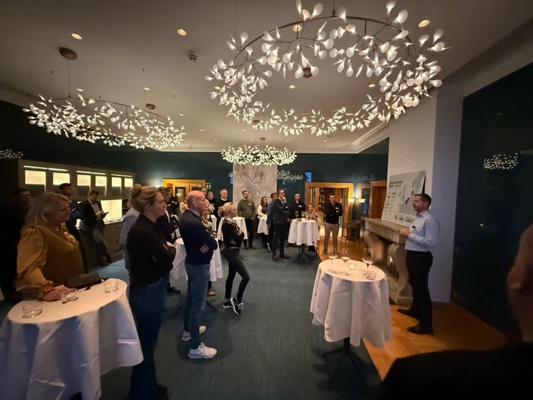 A person is speaking to a group of people gathered in a well-lit room with decorative ceiling lights and standing tables.